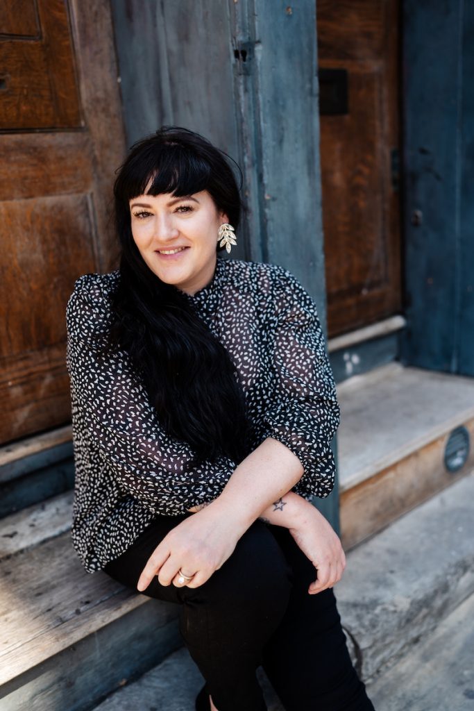 Natalie, wedding photographer, seated on a wooden step in front of weathered timber doors, smiling in soft daylight